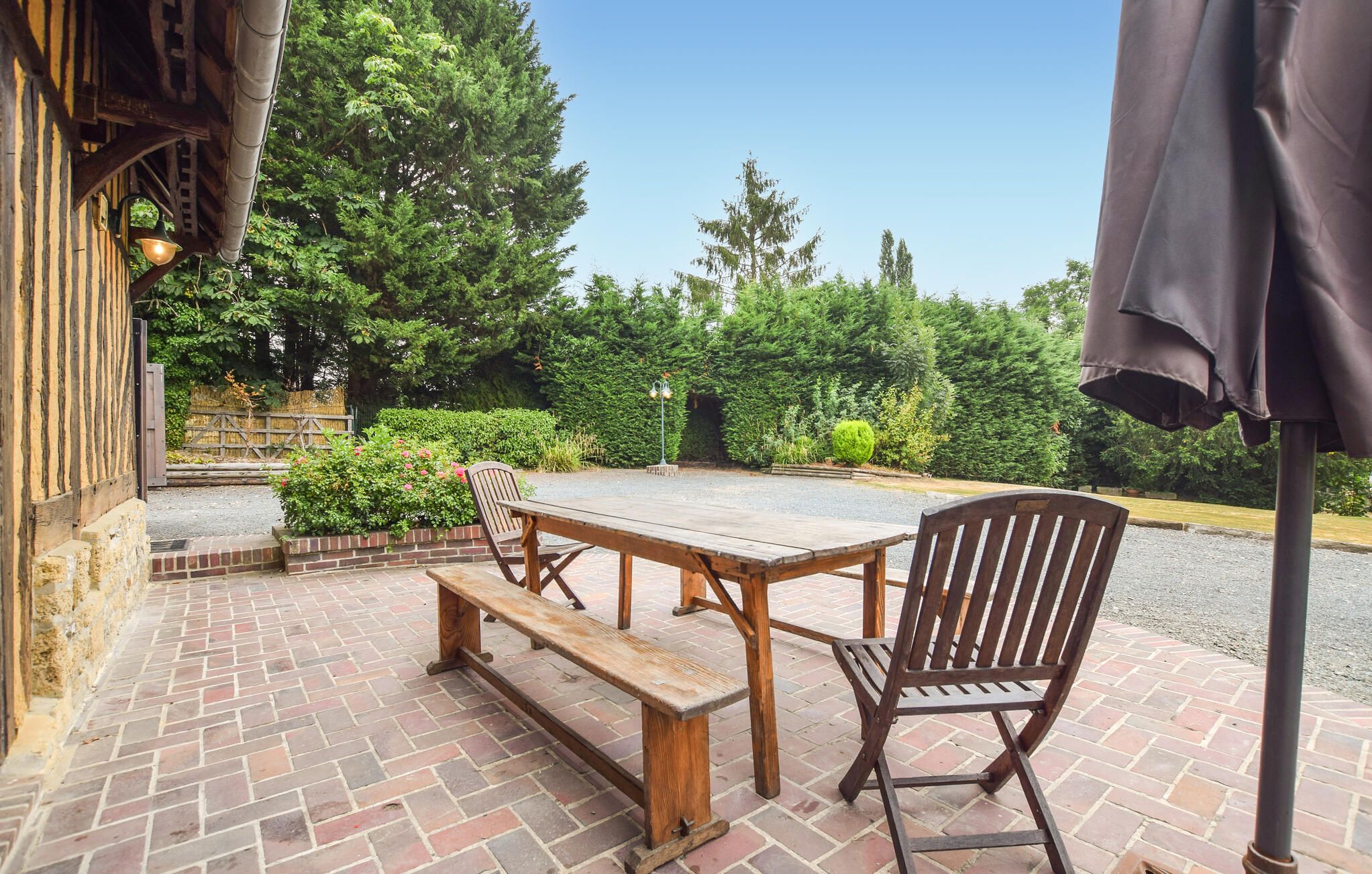 Une terrasse extérieure avec une table et des bancs en bois, entourée de verdure et bordée d’un chemin en briques.