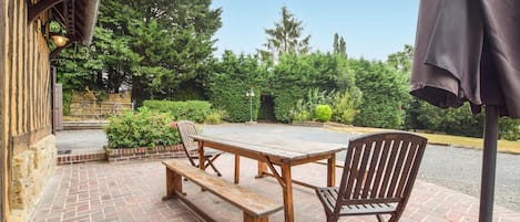 Une terrasse extérieure avec une table et des bancs en bois, entourée de verdure et bordée d’un chemin en briques.