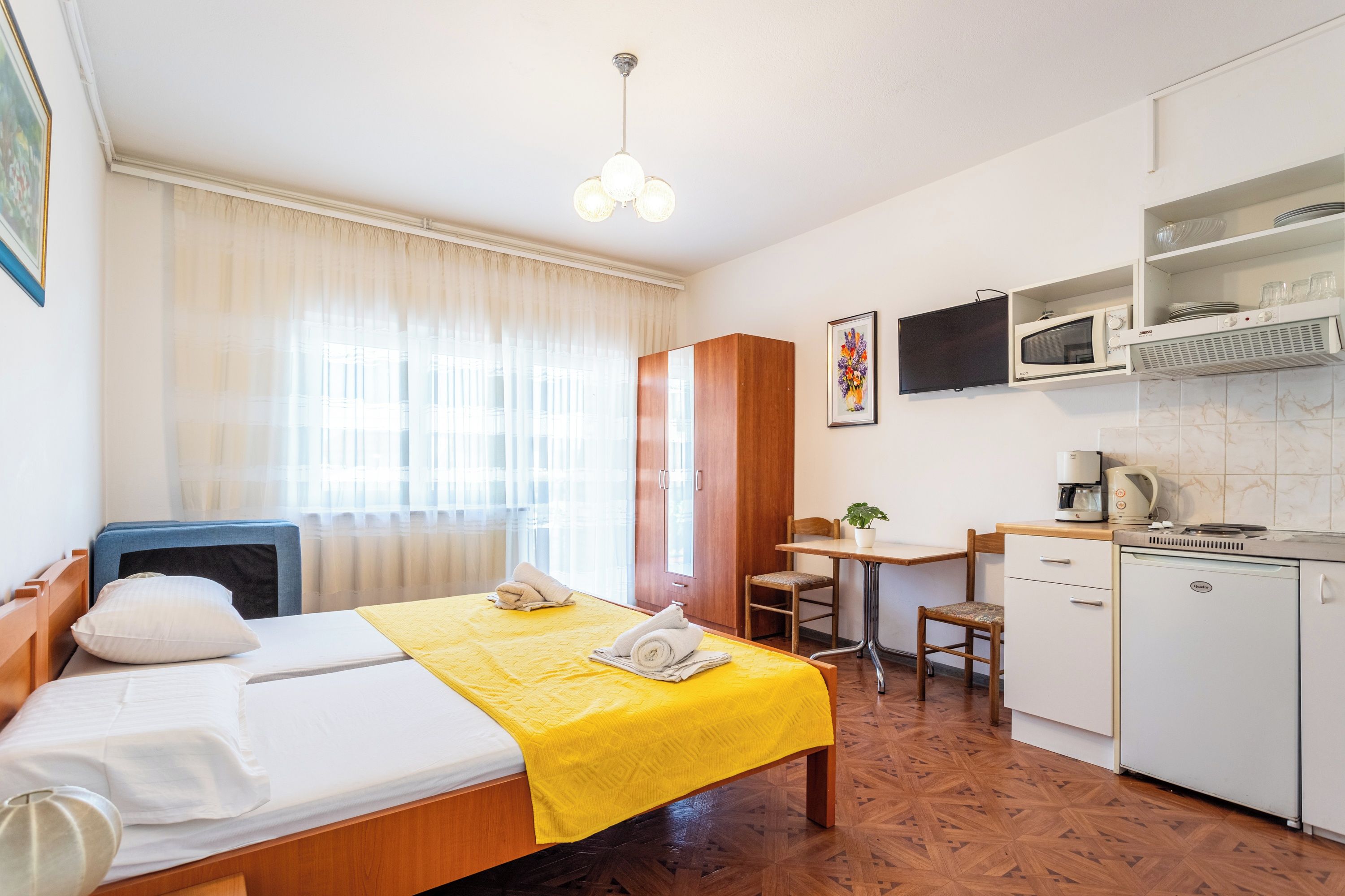 A hotel room with a bed, a desk, a chair, a kitchenette, and a window with curtains.