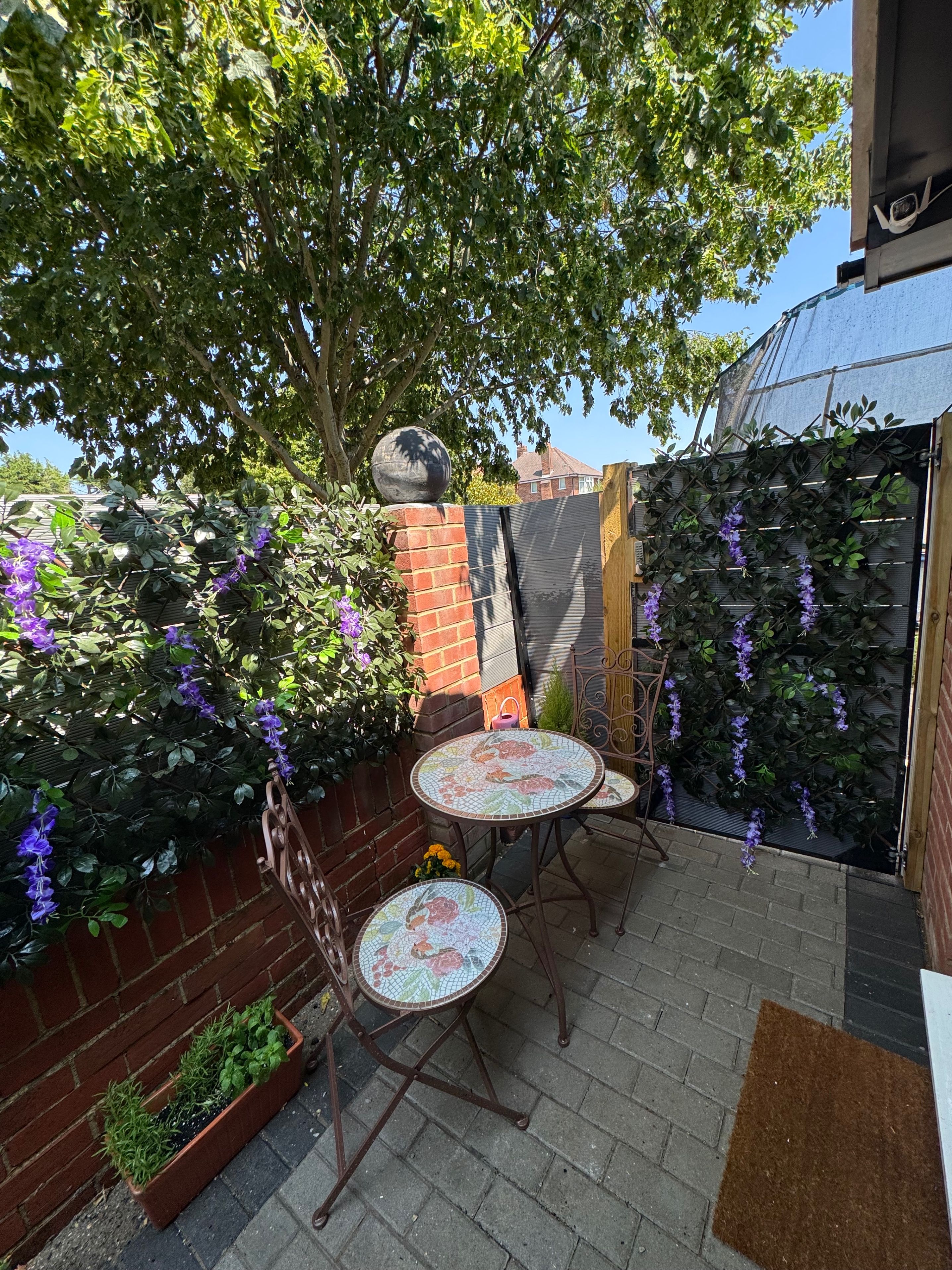 A patio with two mosaic-tiled tables, a brick wall, and a climbing plant with purple flowers.