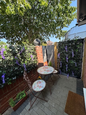 Een terras met twee mozaïek betegelde tafels, een bakstenen muur en een klimplant met paarse bloemen.