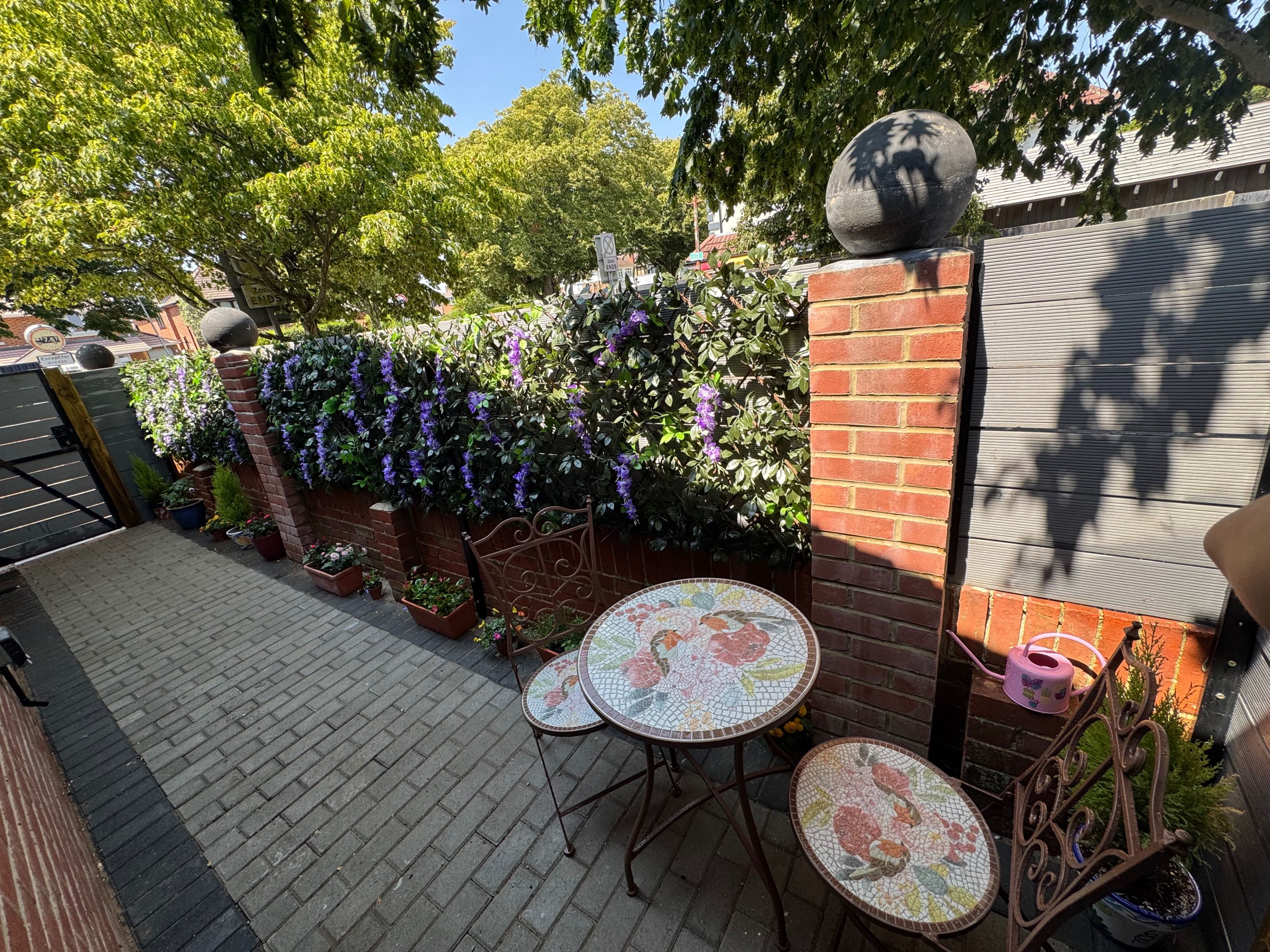 A patio with a tiled table and chairs, surrounded by a brick wall with climbing plants and a metal gate.