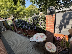 Een terras met een betegelde tafel en stoelen, omringd door een bakstenen muur met klimplanten en een metalen poort.
