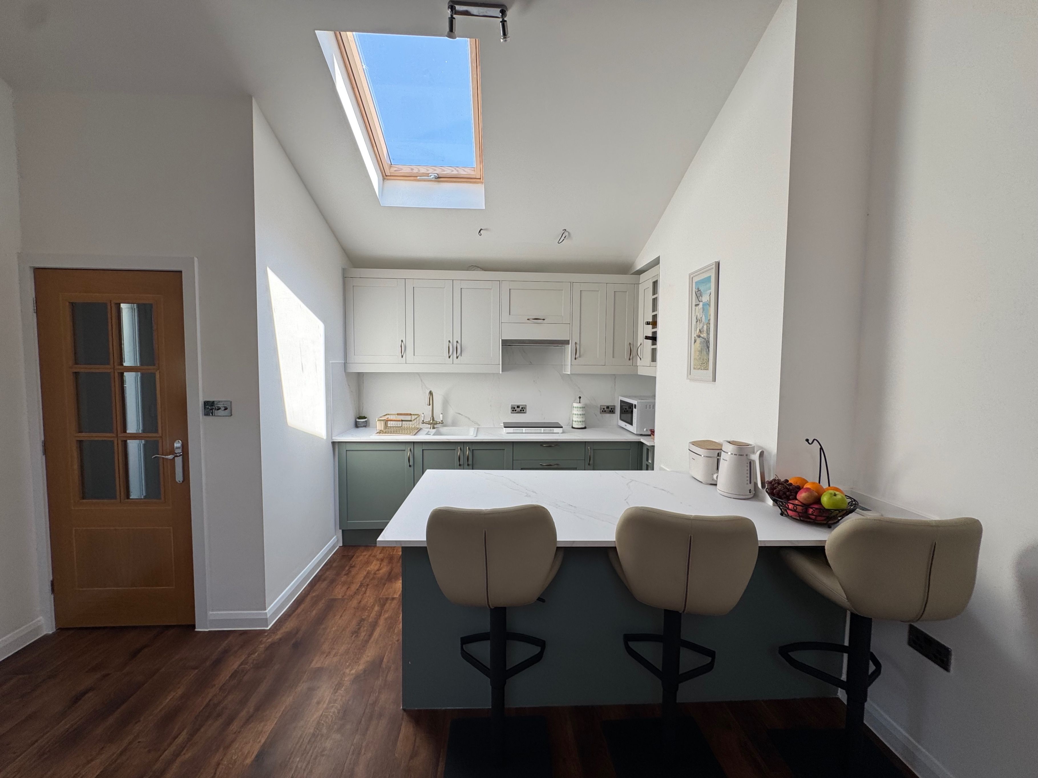 A modern kitchen with a skylight, a wooden door, and a bar area with three stools.