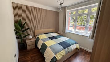 A bedroom with a wooden slat wall, a bed with a checkered bedspread, a nightstand, a potted plant, and a window with a view of the outside.