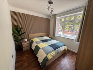 Un dormitorio con pared de listones de madera, una cama con colcha escocesa, una mesita de noche, una planta en maceta y una ventana con vistas al exterior.