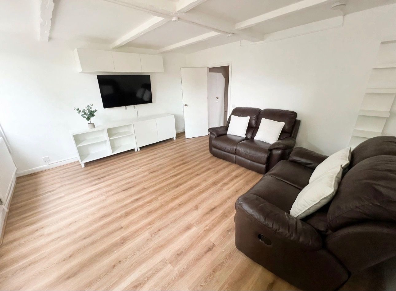 A spacious living room with a brown leather sofa, a flat-screen TV mounted on the wall, and a wooden floor.