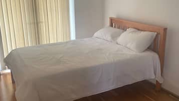 Un dormitori amb un llit de fusta, roba de llit blanca i una finestra amb persianes.