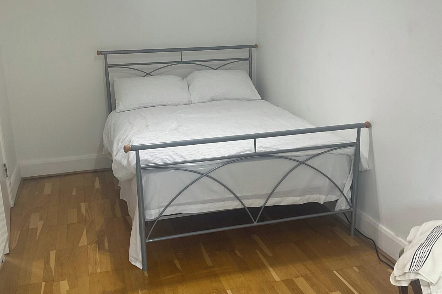 Un llit amb estructura metàl·lica i roba de llit blanca en una habitació amb terres de fusta i parets blanques.