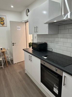 A compact kitchen with white cabinets, a black induction cooktop, a microwave, and a stainless steel range hood. - Private Bedroom with kitchen Access (London)
