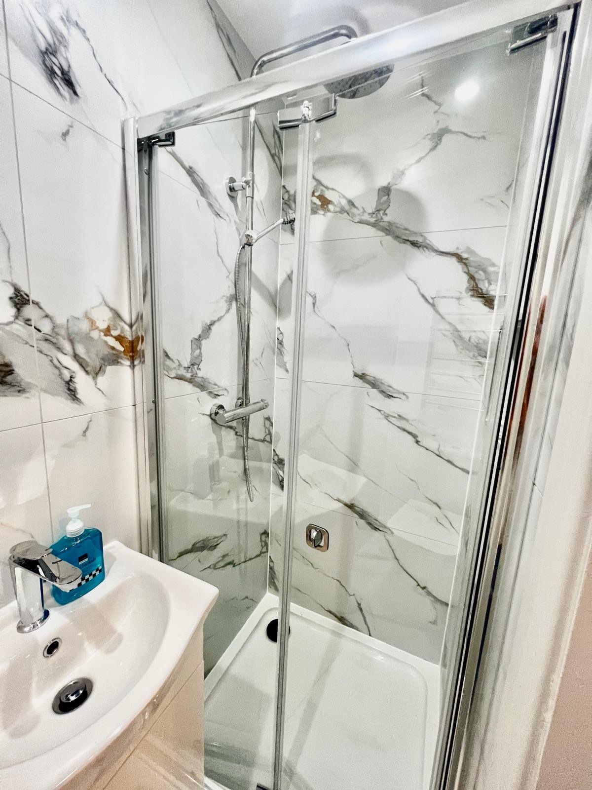 Ein Badezimmer mit einer Duschkabine aus Glas, einem Waschbecken mit Seifenspender und Wänden mit Marmoroptik.