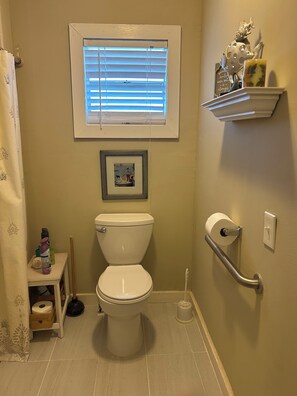 A bathroom with a toilet, a window with blinds, a picture frame, a shelf with toiletries, and a handrail.