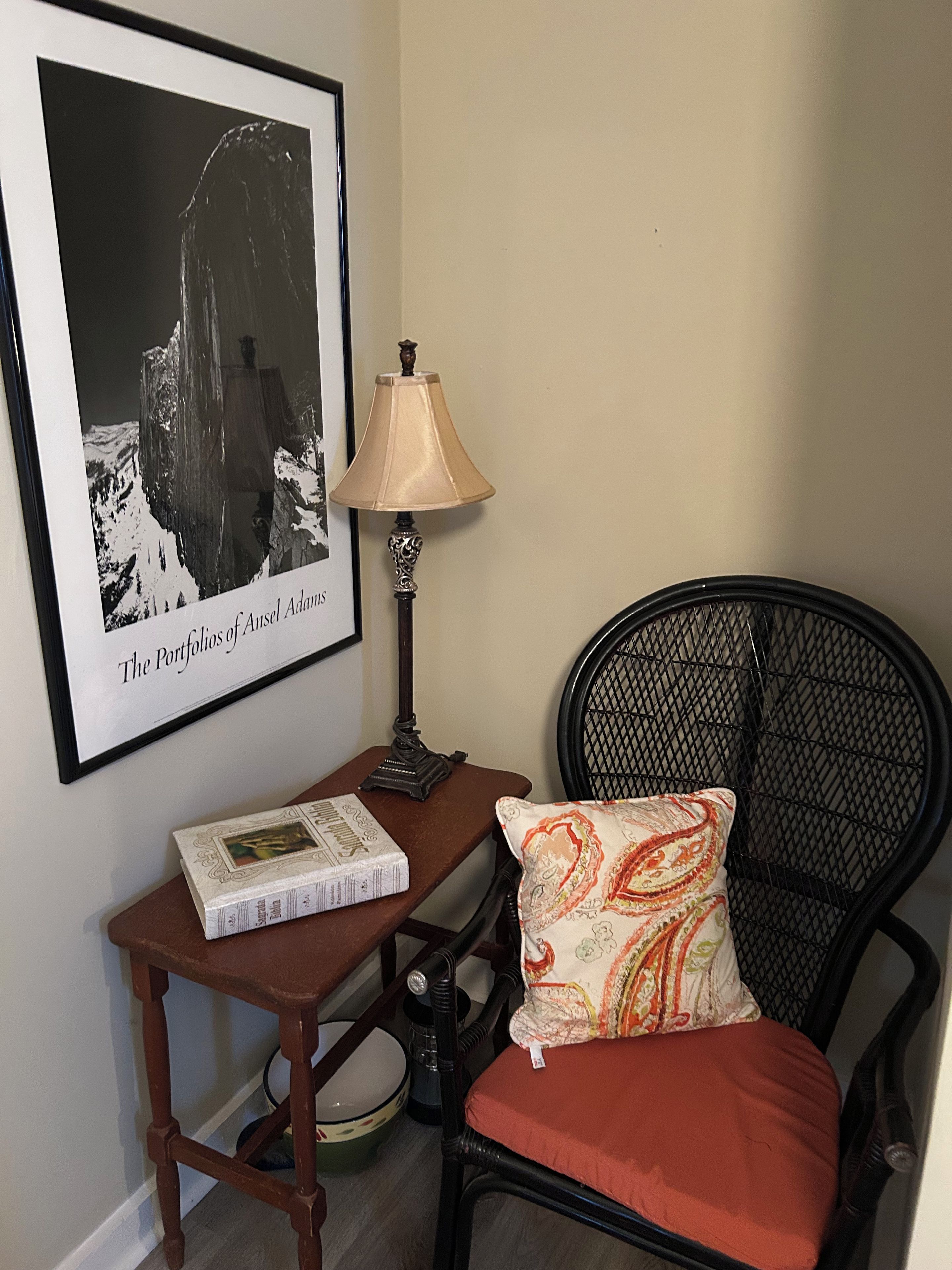 A corner of a room with a framed photograph, a table with a lamp, a chair with a cushion, and a book.