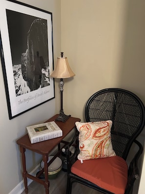 A corner of a room with a framed photograph, a table with a lamp, a chair with a cushion, and a book.