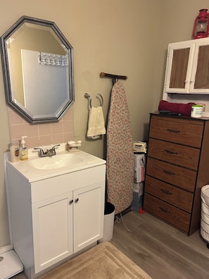 Un bagno con un lavandino bianco, uno specchio, un portascopevoli, un asse da stiro e un armadietto in legno.