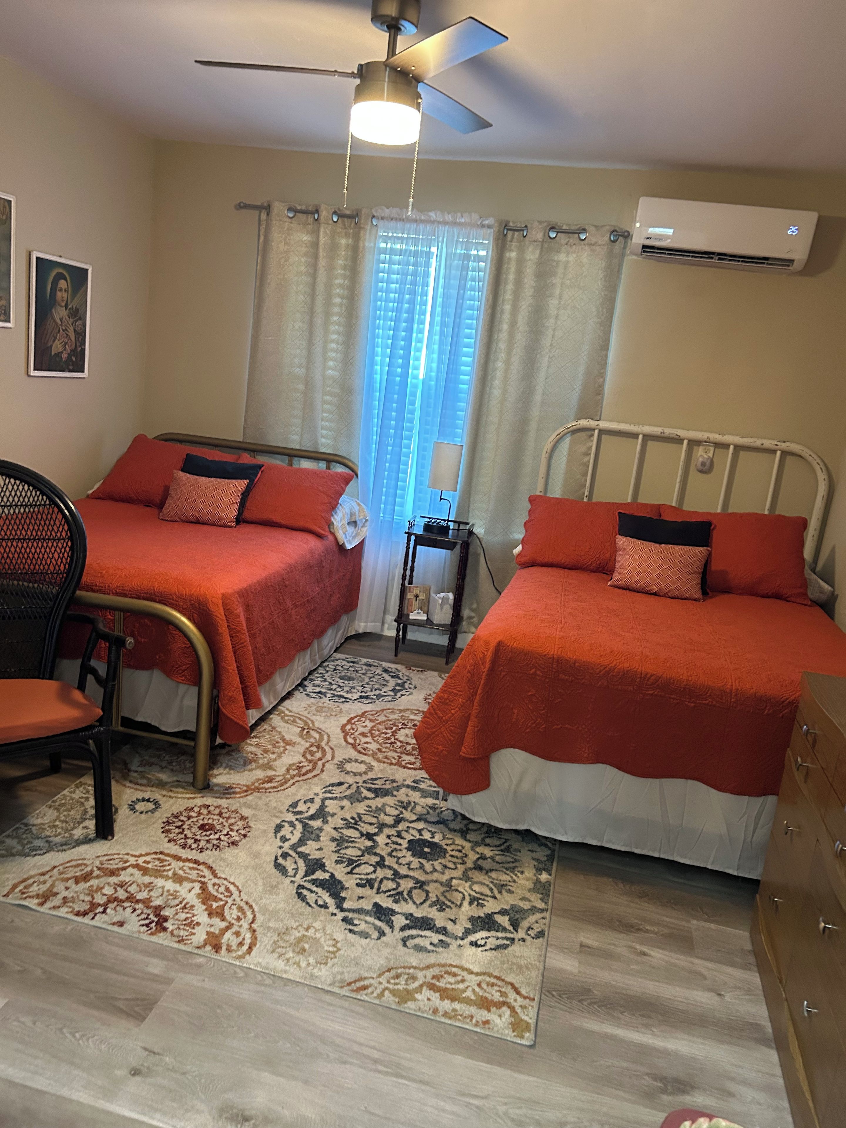 A room with two beds, a ceiling fan, an air conditioning unit, a small table, a chair, and a patterned rug.