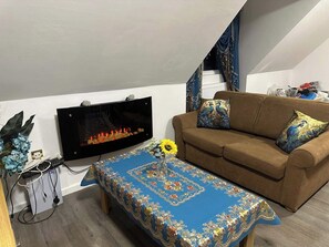 En stue med et fladskærms-tv, en brun sofa med pyntepuder, en blå borddug med mønster og en lille plante.