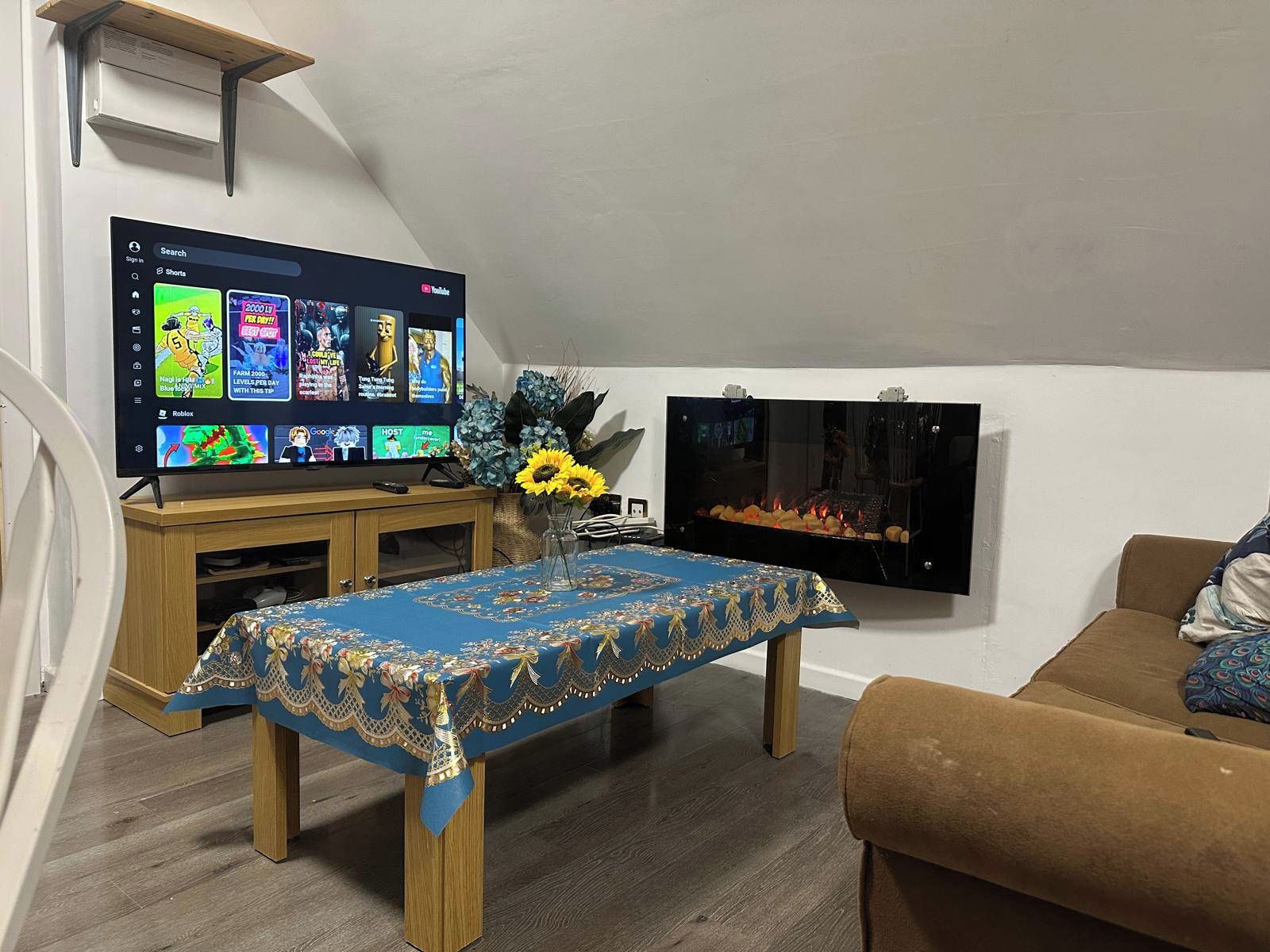 Ein Wohnzimmer mit einem wandmontierten Flachbildfernseher, einem Couchtisch aus Holz mit blauer Tischdecke, einem braunen Sofa und einem kleinen Tisch mit einer Vase voller Sonnenblumen.