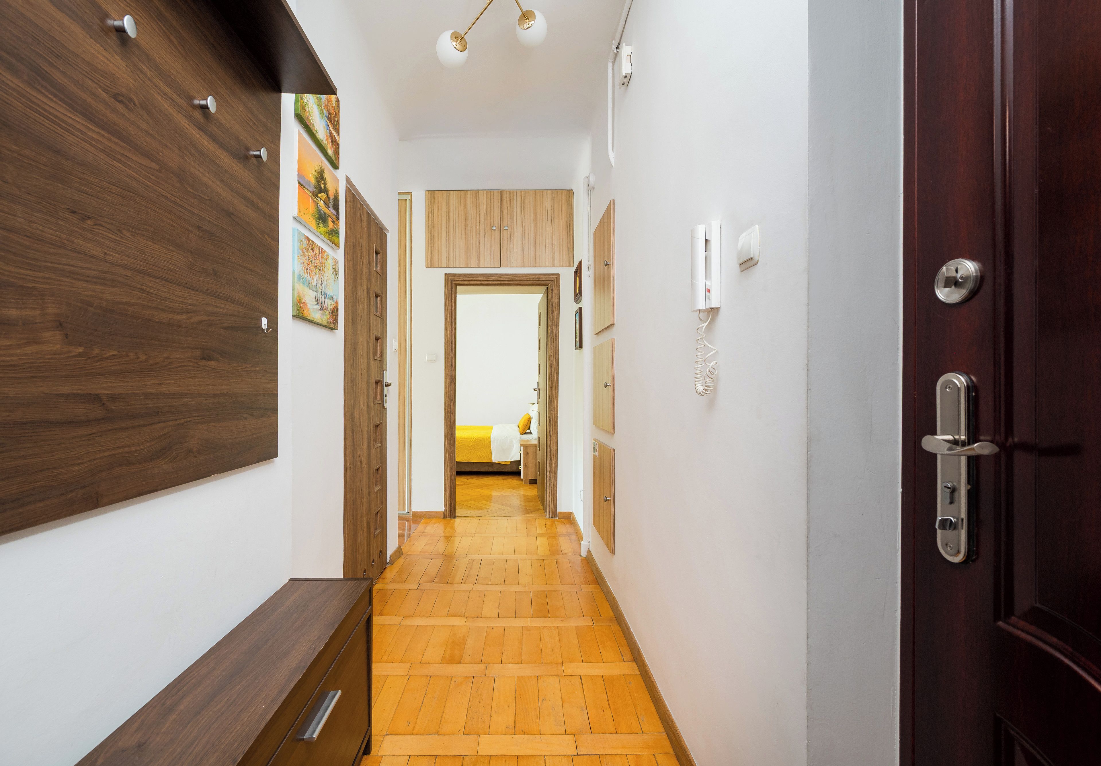 A hallway with wooden flooring, a wooden door on the right, and a bed visible in the distance.