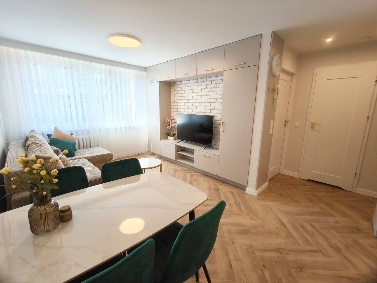 A modern living room with a dining area, a TV mounted on a brick wall, and a large window with curtains.