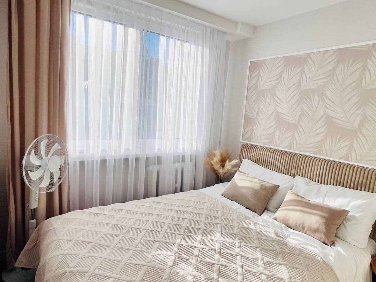 Una habitación de hotel con una cama, un ventilador de pie, cortinas traslúcidas y un cabecero estampado.