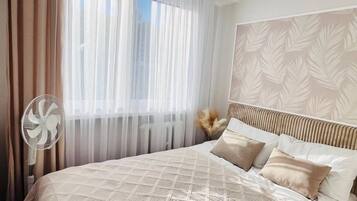 Una habitación de hotel con una cama, un ventilador de pie, cortinas traslúcidas y un cabecero estampado.
