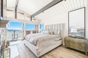 A bedroom with a large bed, a nightstand, a lamp, and a painting. There is a view of the beach through the window. - Coveted Beachfront Home with Oceanfront Patio and Balcony (Newport Beach)