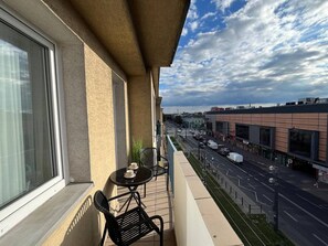 A balcony with a table and chairs, overlooking a street with buildings and parked cars.