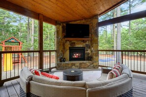 A covered outdoor seating area with a stone fireplace, a children's play structure, and a view of the surrounding forest.