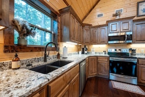 A kitchen with wooden cabinets, granite countertops, stainless steel appliances, and a window with a view of trees.