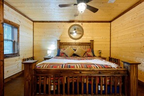A wooden-paneled bedroom with a bed, a ceiling fan, a round mirror, and a window with blinds.