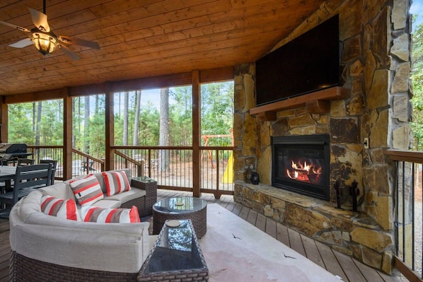 A covered outdoor area with a stone fireplace, seating, and a view of the surrounding forest.
