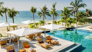 Área de piscina em resort com espreguiçadeiras e guarda-sóis, cercada por palmeiras e vista para o oceano.