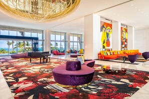 A spacious lobby with a grand chandelier, large windows offering a view of the ocean, and vibrant artwork on the walls.