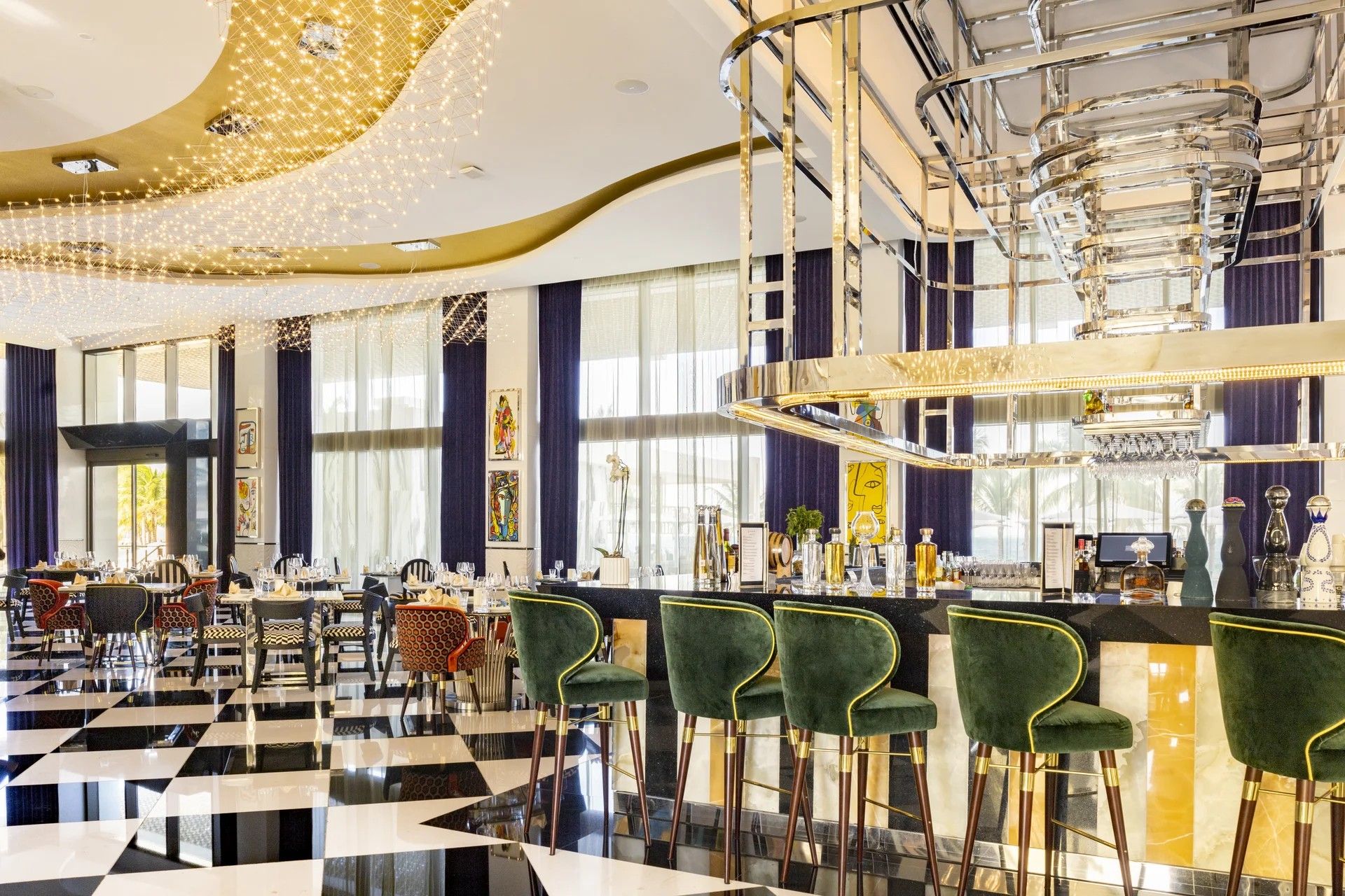 Een bar met groene bekleden barkrukken, een geruit vloertje en een moderne kroonluchter.