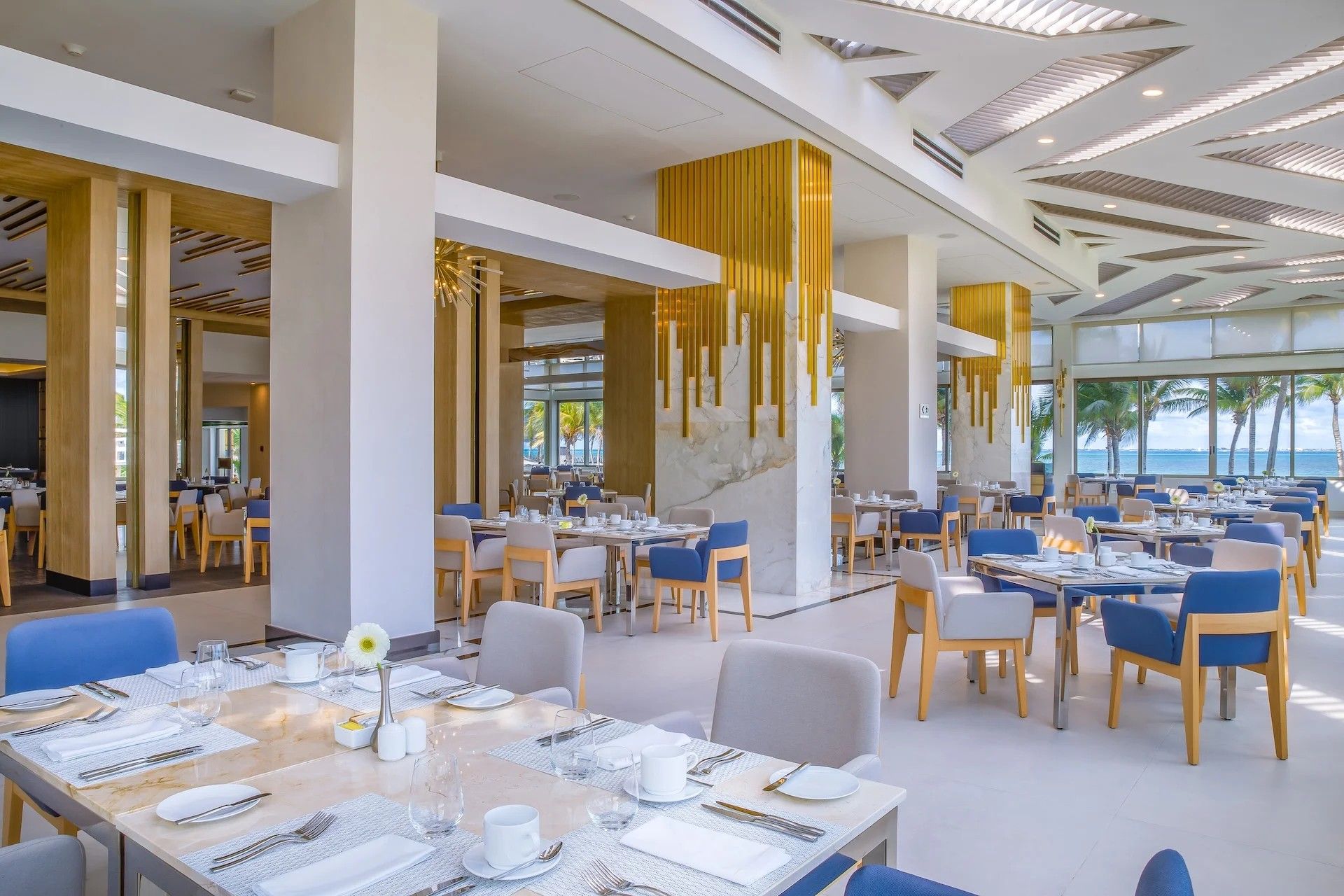 A modern dining area with multiple tables and chairs, a view of the ocean, and a ceiling with unique lighting fixtures.