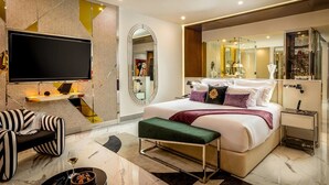 Quarto de hotel moderno com cama grande, TV de tela plana, área de estar com sofá e poltrona, e banheiro com espelho amplo.