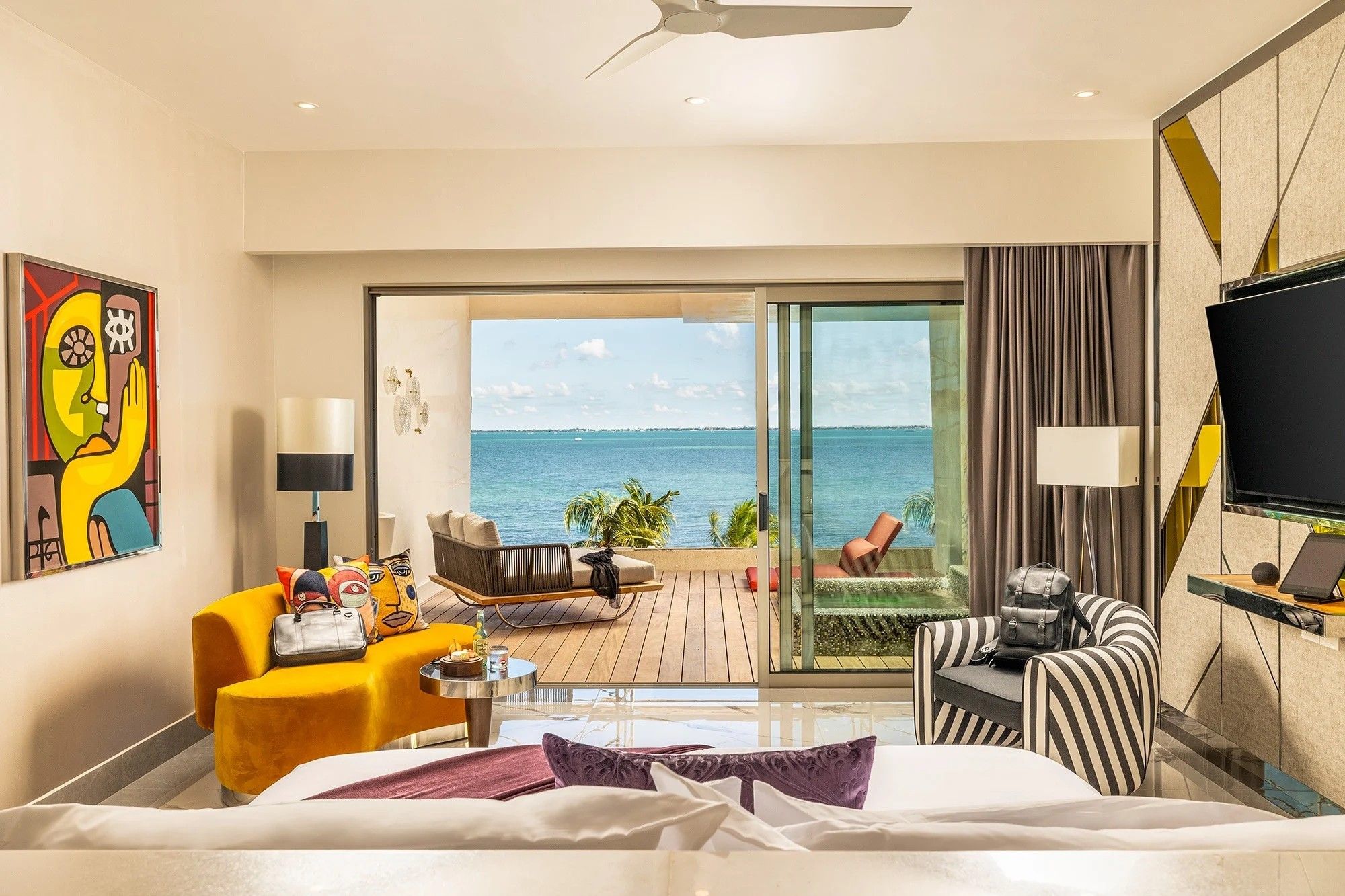 A hotel room with a bed, a sofa, a chair, a small table, a TV, and a balcony with a view of the sea.