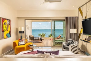 A hotel room with a bed, a sofa, a chair, a small table, a TV, and a balcony with a view of the sea.