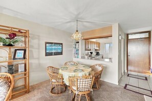 A dining area with a round table, wicker chairs, a hanging light fixture, and a bookshelf with decorative items. - Menehune Shores #321 - Oceanview 2BR Condo, Private Lanai & Steps to the Beach (Kihei)