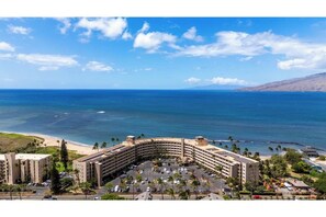 A hotel with a curved building, parking lot, and beachfront view. - Menehune Shores #321 - Oceanview 2BR Condo, Private Lanai & Steps to the Beach (Kihei)