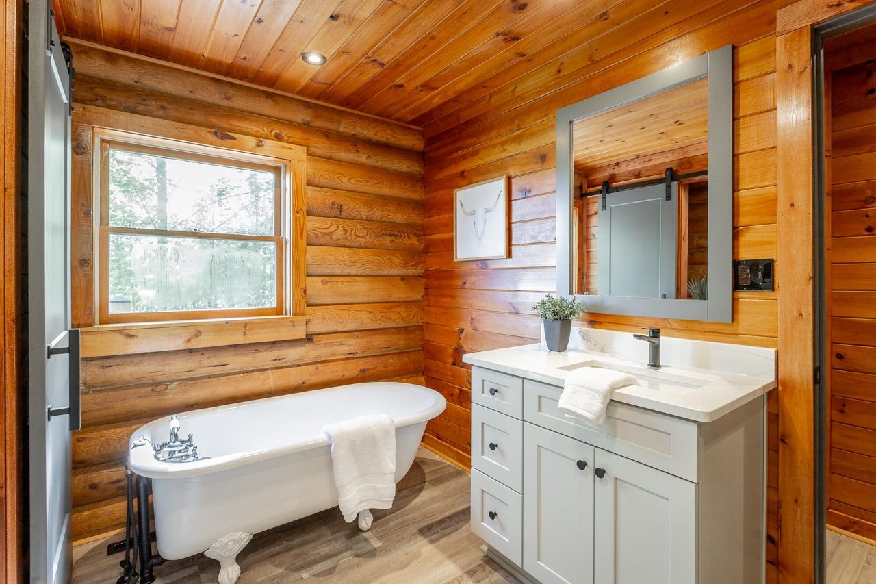 Une salle de bain rustique, avec des murs en bois, comprenant une baignoire sur pieds, un lavabo avec un miroir et une fenêtre laissant entrer la lumière naturelle.