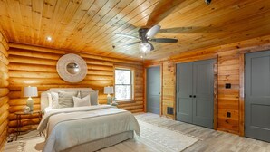 Un dormitorio tipo cabaña de troncos con una cama, ventilador de techo y un ventanal grande.