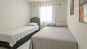 Une chambre avec deux lits simples, une tête de lit, une fenêtre avec des rideaux et un tableau au mur.