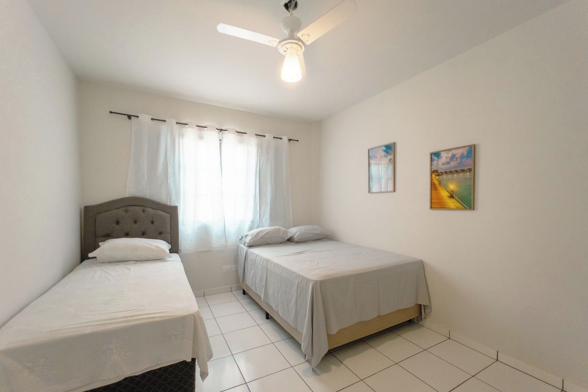 Une chambre avec deux lits simples, un ventilateur de plafond et deux tableaux accrochés au mur.