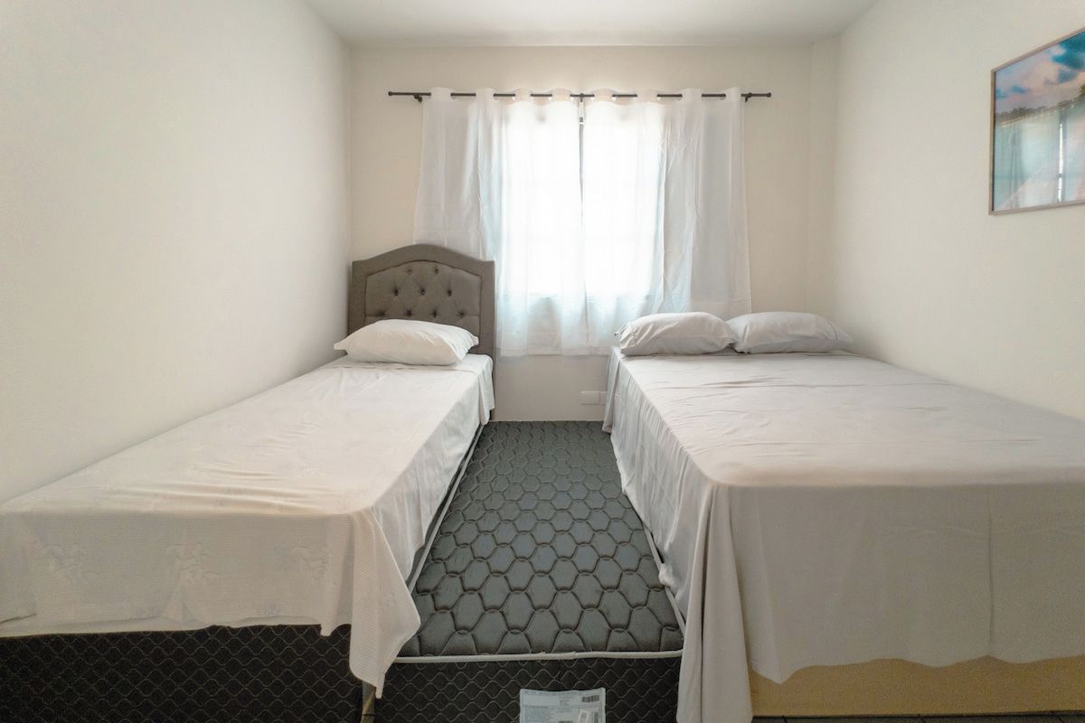 Une petite chambre d’hôtel composée d’une seule pièce, avec deux lits simples, une fenêtre avec des rideaux et une vue sur une étendue d’eau.