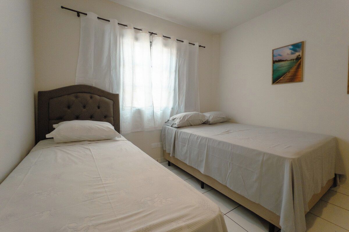 Un dormitorio con dos camas, cabecero de madera, ropa de cama blanca, una ventana con cortinas blancas y un cuadro en la pared.