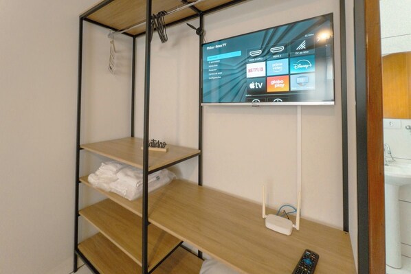 A hotel room with a mounted TV displaying various streaming services, a wooden shelf unit, and a remote control on the floor. - Budget Apartment in Zona 7 Maringa - AL09 (Maringá)
