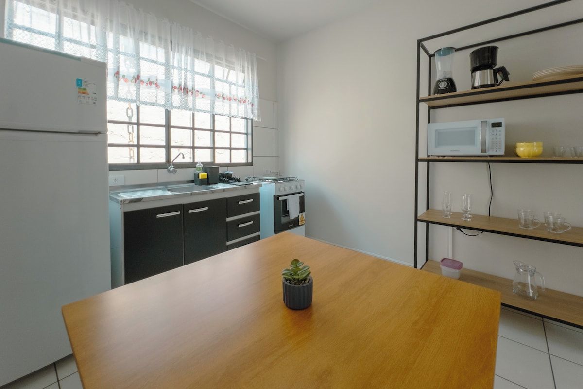 A kitchen with a wooden table, a refrigerator, a microwave, and a shelf with kitchenware.