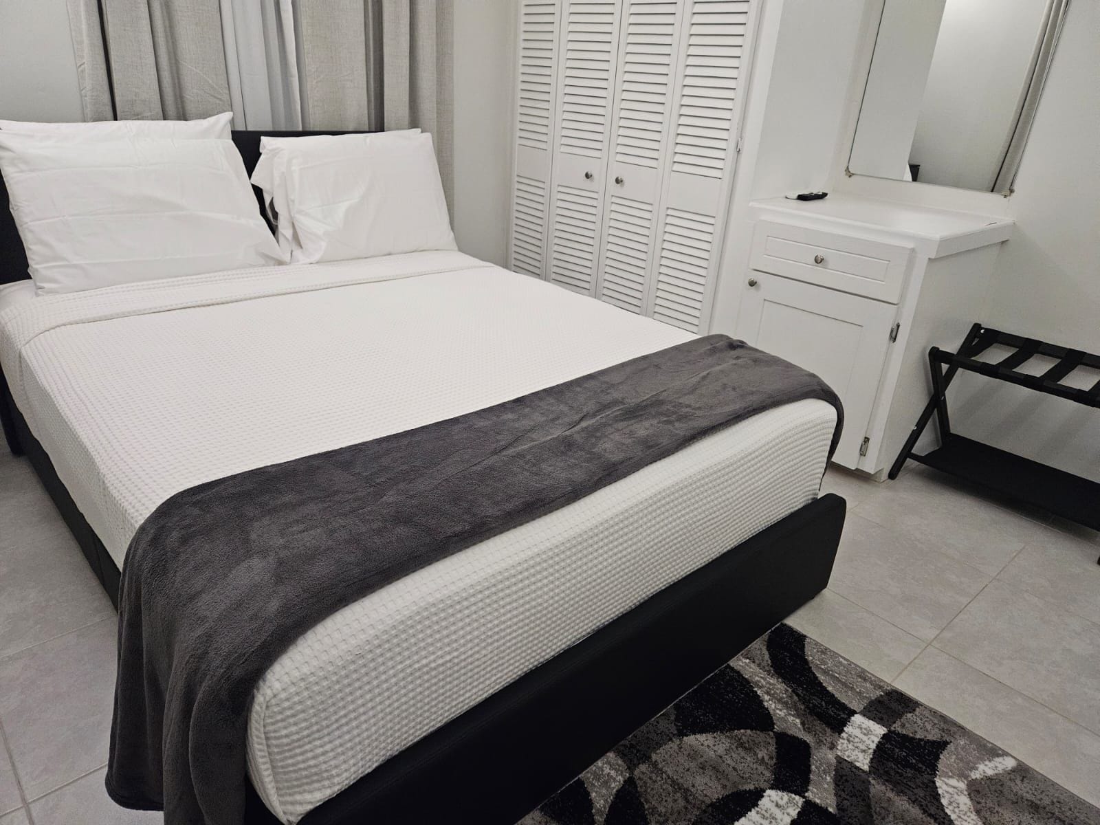Un lit bien fait, avec un matelas blanc et une couverture grise, une table de chevet blanche avec un miroir, et un banc noir.